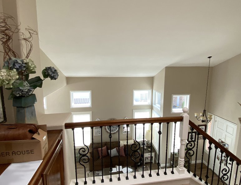 Before photo showing  Upper landing with wood floor and wrought‑iron railing overlooking a living room with small windows, chandelier, and beige walls.