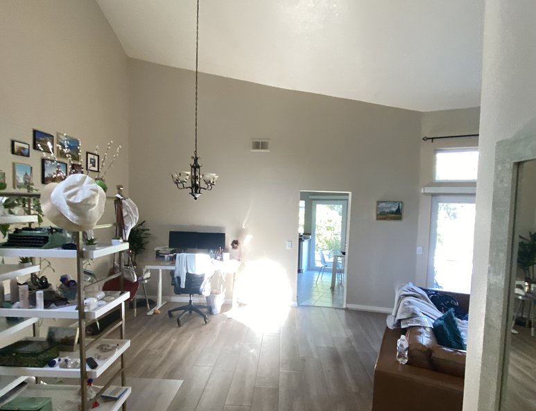 Before photo showing Open living space with vaulted ceilings, wood flooring, a desk setup, shelving unit, chandelier, and a seating area near a sunlit doorway.