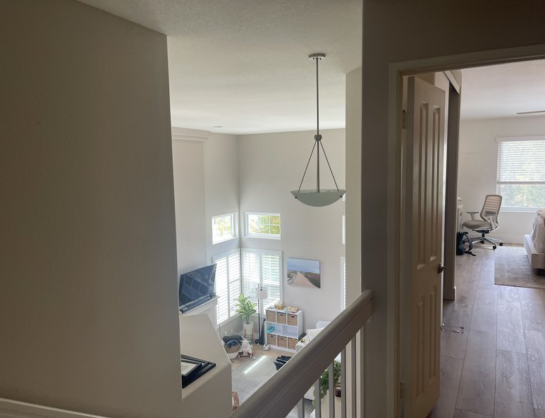 Upper landing before renovation with white railing, wood-look flooring, pendant light, and open overlook into a two-story living space.