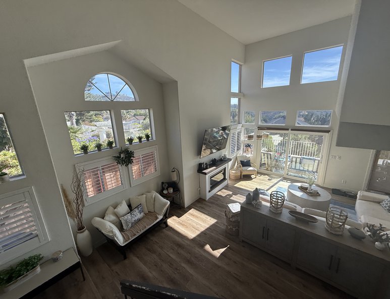 Two-story living room before renovation with tall windows, fireplace, high ceiling, and natural light filling the space.