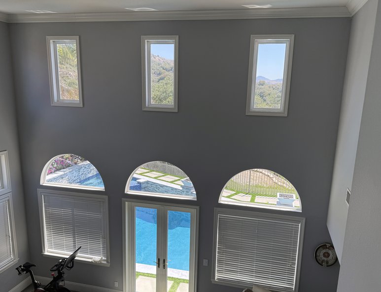 Two-story foyer before renovation with blue front doors, multiple arched and rectangular windows, tall gray walls, and abundant natural light.