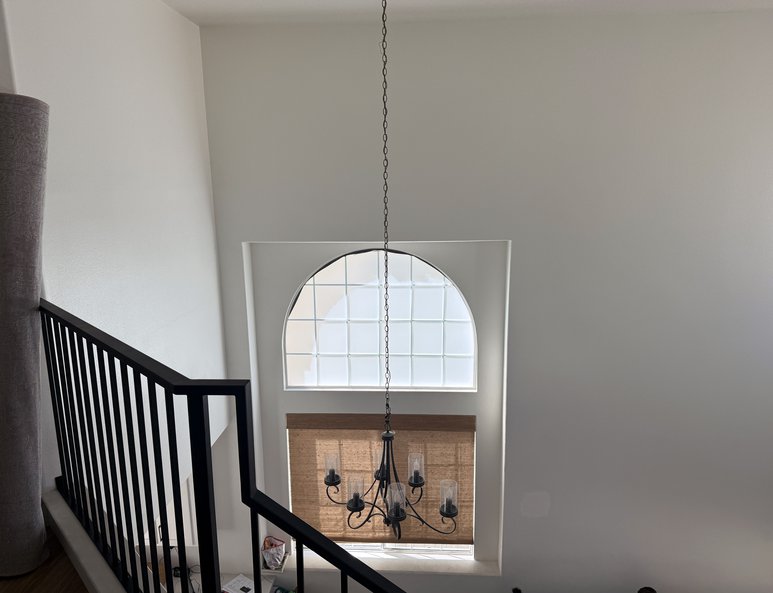 Foyer before renovation with staircase railing, chandelier, arched front-entry window, and two-story ceiling extending above.